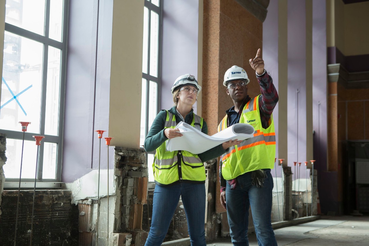 Student and manager in safety vests on a construction site looking at plans and pointing.